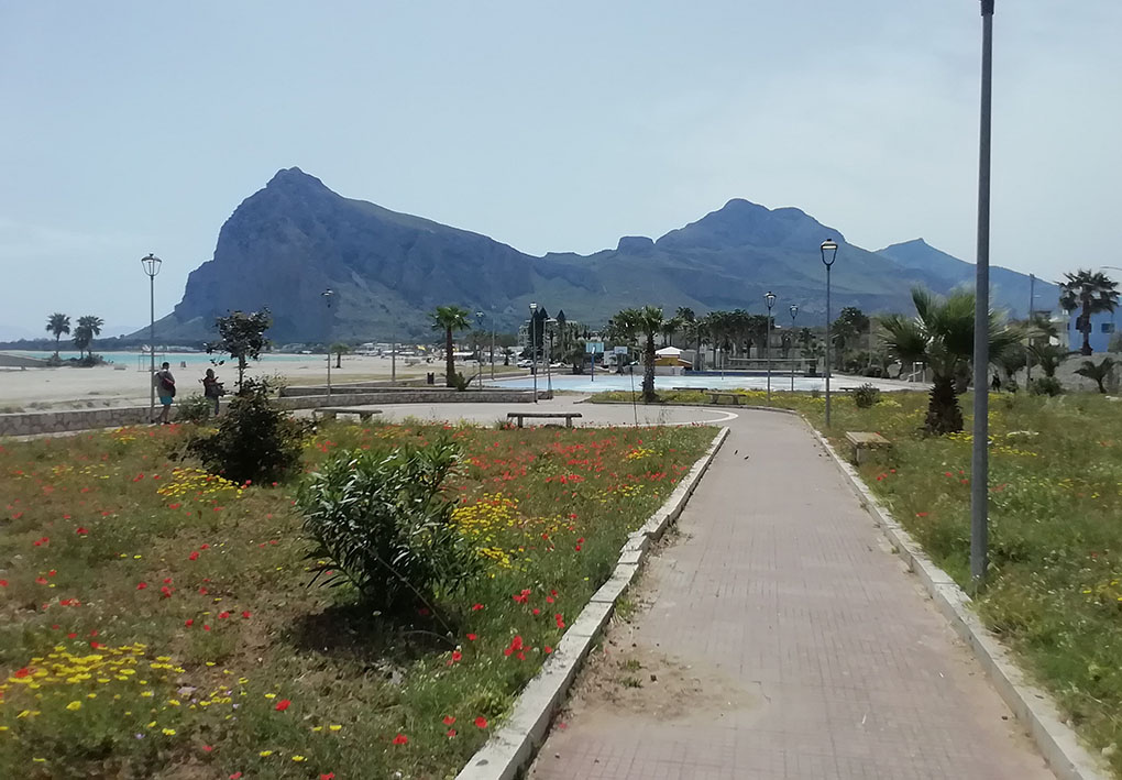 una strada in mezzo ad aiuole, vicina alla costa. Sullo sfondo campi sportivi e un monte in lontananza