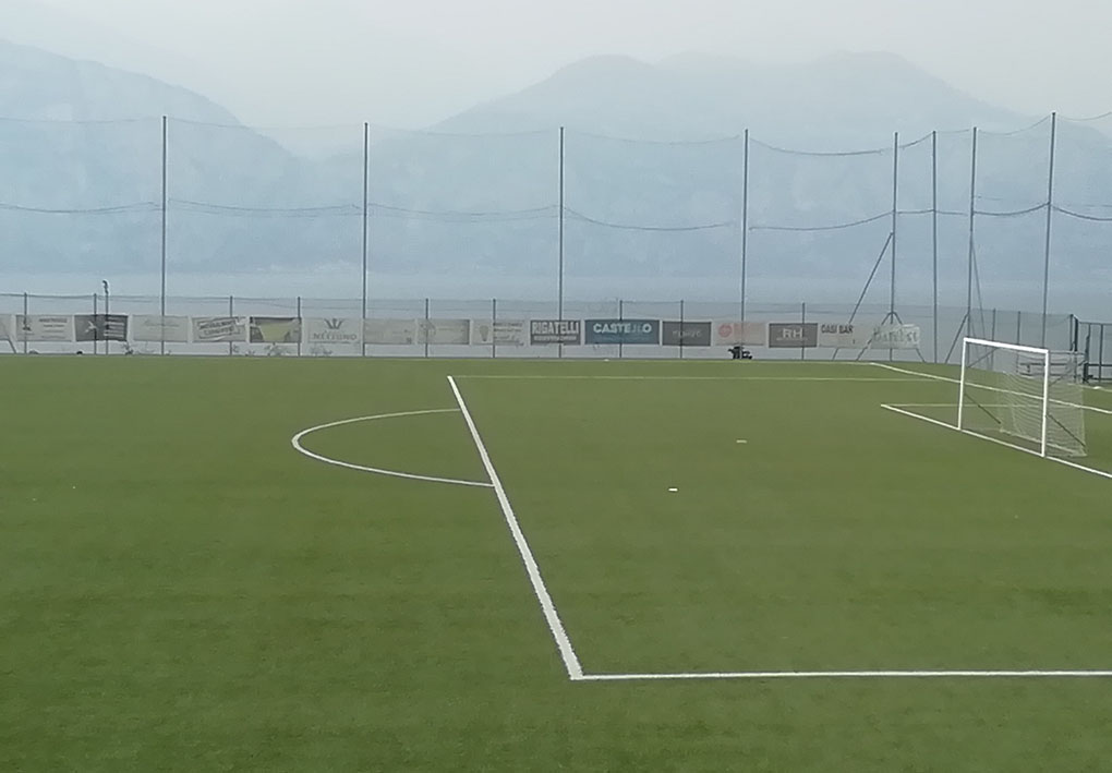 un campo da calcio in erba sintetica, sulle rive del lago. Alte reti e striscioni a bordocampo lo circondano