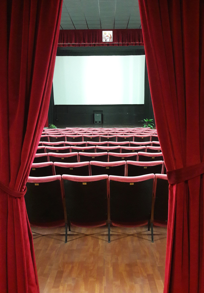 Una sala di un cinema vista dal suo ingresso. Tende di velluto sono tirate ai lati, all'interno si vede la platea di poltroncine da Cinema, rivolte verso uno schermo. Il pavimento è in parquet.