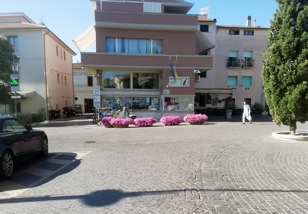 un palazzetto moderno e basso si affaccia su una piazza in sampietrini, con dei cipressi al centro. Fiorirere rigogliose adornano l'ingresso dell'infopoint