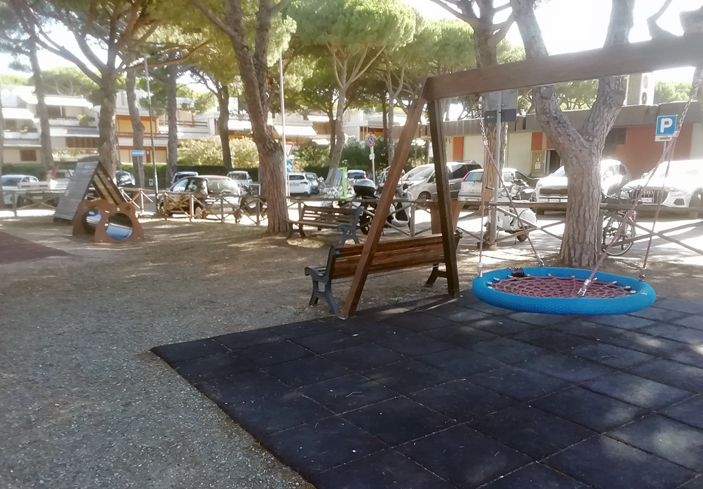 in primo piano una altalena inclusiva tra i pini. Sullo sfondo altri giochi in legno, ogni area gioco è provvista di piastrelloni antitrauma