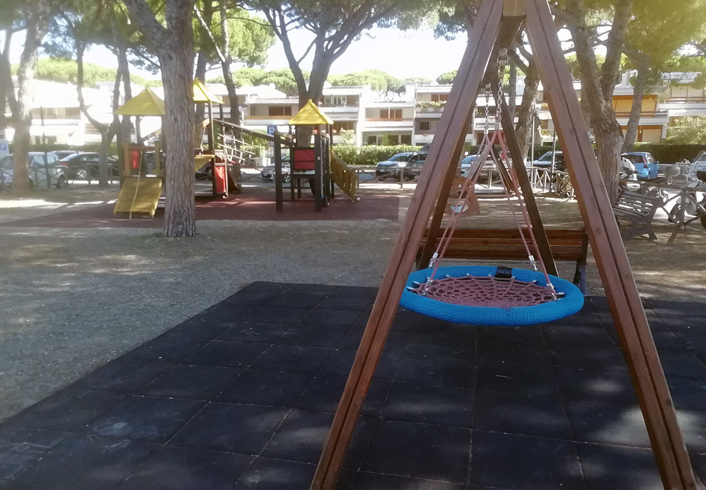 in primo piano una altalena inclusiva tra i pini. Sullo sfondo altri giochi in legno, ogni area gioco è provvista di piastrelloni antitrauma
