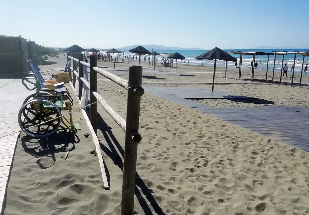 una spiaggia di sabbia punteggiata di ombrelloni e gazebi, percorsa da pedane di legno. Tra le cabine e la spiaggia, sedie JOB sono a disposizione degli utenti che ne hanno bisogno.