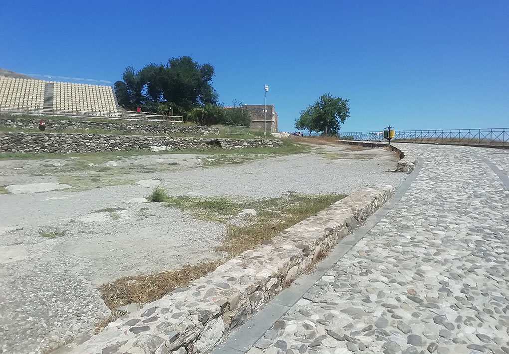 Percorso su antiche mura che si intuisce si affacciano sul mare. Sul lato, una platea di sedie in plastica di un'area spettacoli