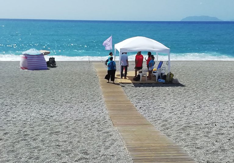 una pedana in legno si allunga verso la spiaggia, dove un gazebo con la bandiera lilla crea un'area ombreggiata