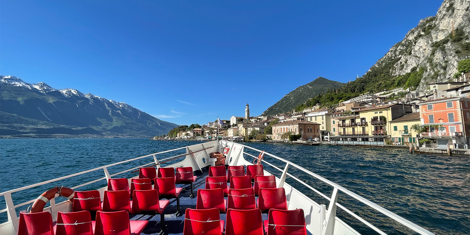 Inquadrata dal ponte aperto, la prua di una nave con tanti seggiolini si dirige verso un borgo sulle rive del lago. Alle spalle del centro abitato si alzano montagne rocciose coperte di vegetazione, sotto a un cielo sereno
