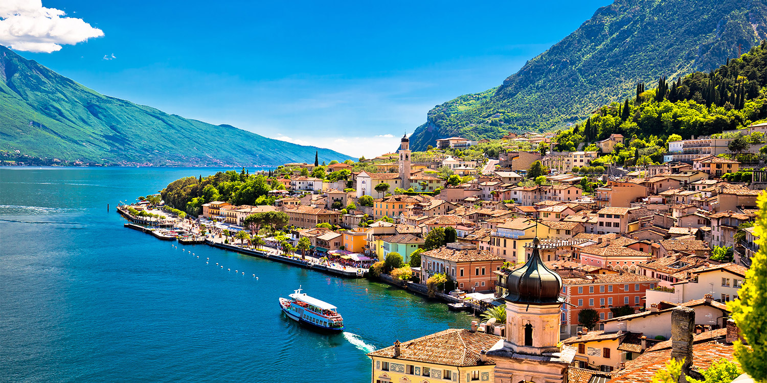 Ripreso dal un'altura, un traghetto si allontana da un varioponto borgo che si affaccia sulle rive del lago. Il cielo è sereno e tutto intorno si alzano alte montagne coperte di vegetazione