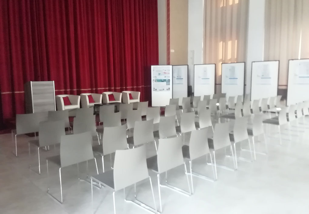 un'ampia sala con cartelli espositivi ai lati. Al centro sedie di plastica rimovibili formano una platea di fronte a un sipario chiuso