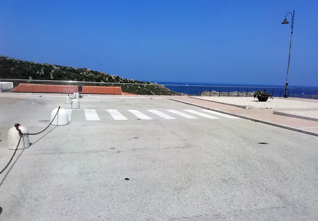Un'area di parcheggio è recitantata da panettoni di cemento e catene. All'uscita di questa are sono segnate chiaramente le strisce pedonali che attraversano la carreggiata. Dall'altro lato, il marciapiede pedonale. Sullo sfondo una ringhiera sulla costa sottostante
