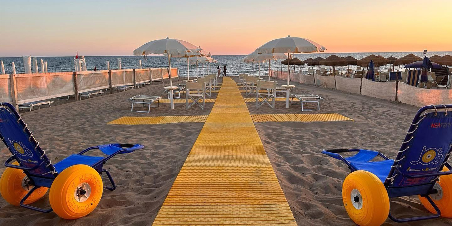 La spiaggia al tramonto. In primo piano si trovano due sedie job, mentre la passerella si allunga verso il mare tra file ordinate di ombrelloni