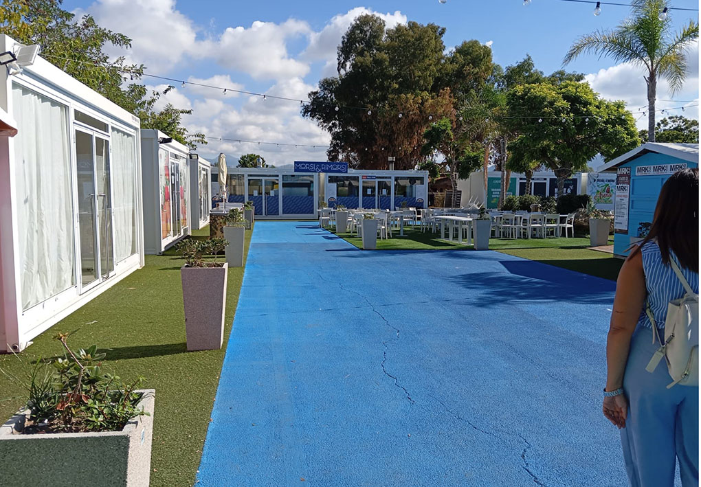 Cortile interno con una passatoia in asfalto per accedere alla zona centrale con i tavoli e gli alberi. La zona è circondata da basse costruzioni moderne, una delle quali sembra essere un ristorante