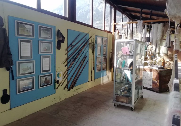 Stanza espositiva con pannelli contenenti fotografie e testi informativi. Tra i pannelli sono appesi indumenti, fucili e attrezzi da pesca. Al centro, una vetrina su ruote mostra oggetti vari, tra cui libri e strumenti. Sullo sfondo, reti da pesca appese e una statua arricchiscono l’ambiente. Grandi finestre permettono di vedere le montagne all’esterno.