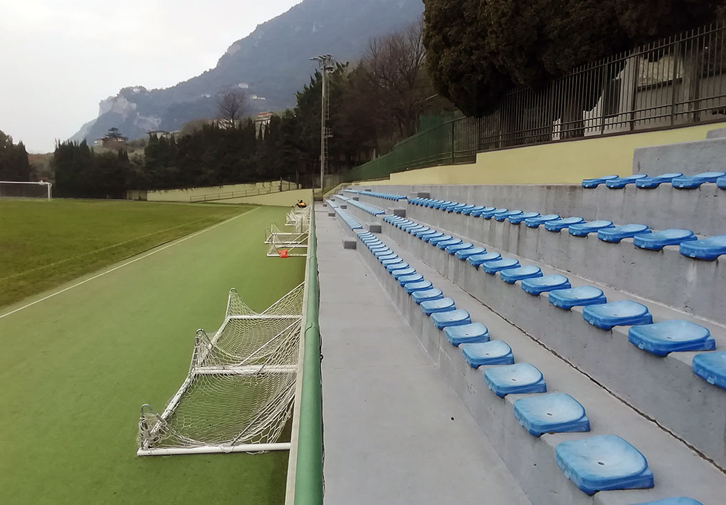 tre file di spalti con seggiolini in plastica affiancano il lato lungo del campo da calcio. In cima agli spalti prosegue la vegetazione che circonda lo spazio