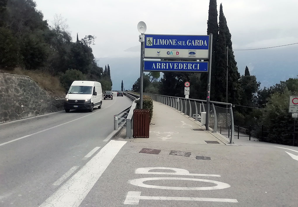 Strada asfaltata in una zona collinare con vegetazione verde. Sul lato sinistro, un muro di pietra segue il percorso della strada, mentre a destra un marciapiede con ringhiera metallica offre una vista aperta sul paesaggio. All'ingresso del percorso, cartelli segnalano l'uscita dal Comune.