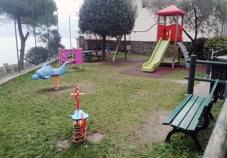 Parco giochi all’aperto con diverse strutture colorate per bambini. Il parco è delimitato da una recinzione e circondato da alberi. Sul lato destro dell’immagine, ci sono due panchine verdi e sullo sfondo si nota un tavolo da picnic. Il prato verde e le aree con tappeti di gomma assicurano uno spazio sicuro e accogliente per il gioco.