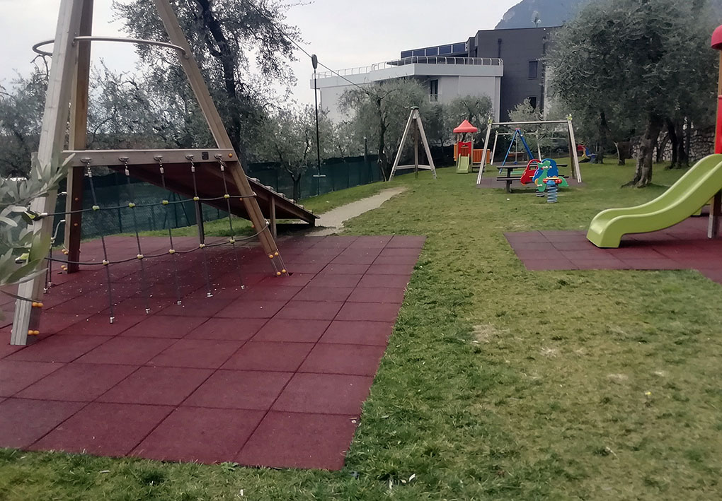 In primo piano, una struttura per teleferica-gioco in legno con rampa di risalita e corde per arrampicarsi. In fondo al parco si nota la corrispettiva struttura di arrivo. Il prato è punteggiato di altri giochi, circondati da piastrelloni antitrauma.