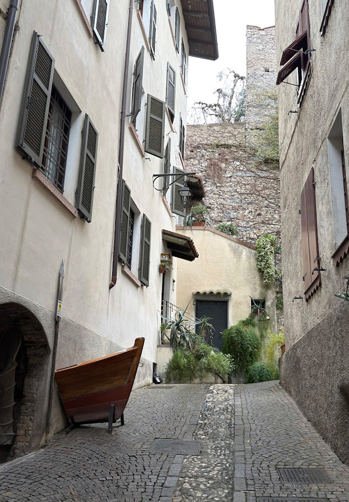 Un vicolo con pavimentazione in sampietrini e piastrelle in salita tra gli edifici. Sulla sinistra, appoggiata al muro di un edificio, è esposta una barca da pesca in legno lucido. Le case hanno facciate chiare con persiane in legno verdi e marroni, sullo sfondo si intravedono piante e vegetazione rampicante, e in alto un antico muro in pietra che sale verso la collina.
