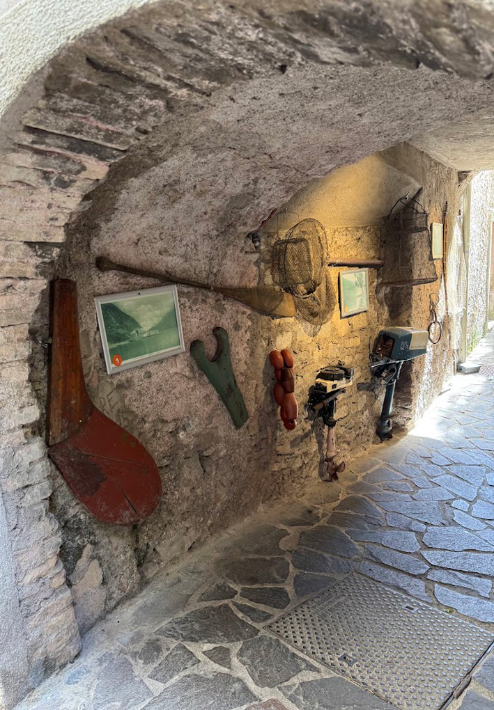 Passaggio ad arco in pietra e mattoni, allestito con oggetti tradizionali da pesca. Sulla parete in pietra sono appesi vecchi remi, reti da pesca, due motori fuoribordo d'epoca, galleggianti e alcune fotografie incorniciate del lago.