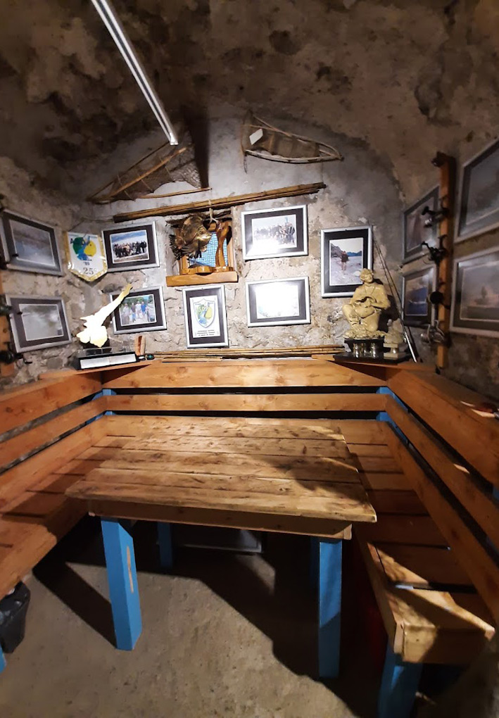 Piccola sala interna in pietra dall'aspetto rustico, arredata come luogo di ritrovo dei pescatori. Al centro un tavolo in legno con gambe azzurre, circondato da panche in legno fissate lungo le pareti. Sulla parete di fondo sono appese numerose fotografie incorniciate, targhe, un gagliardetto e alcuni trofei. In alto sono esposte due tradizionali nasse o attrezzi da pesca in vimini. Sulla destra si intravedono canne da pesca e altri quadri.