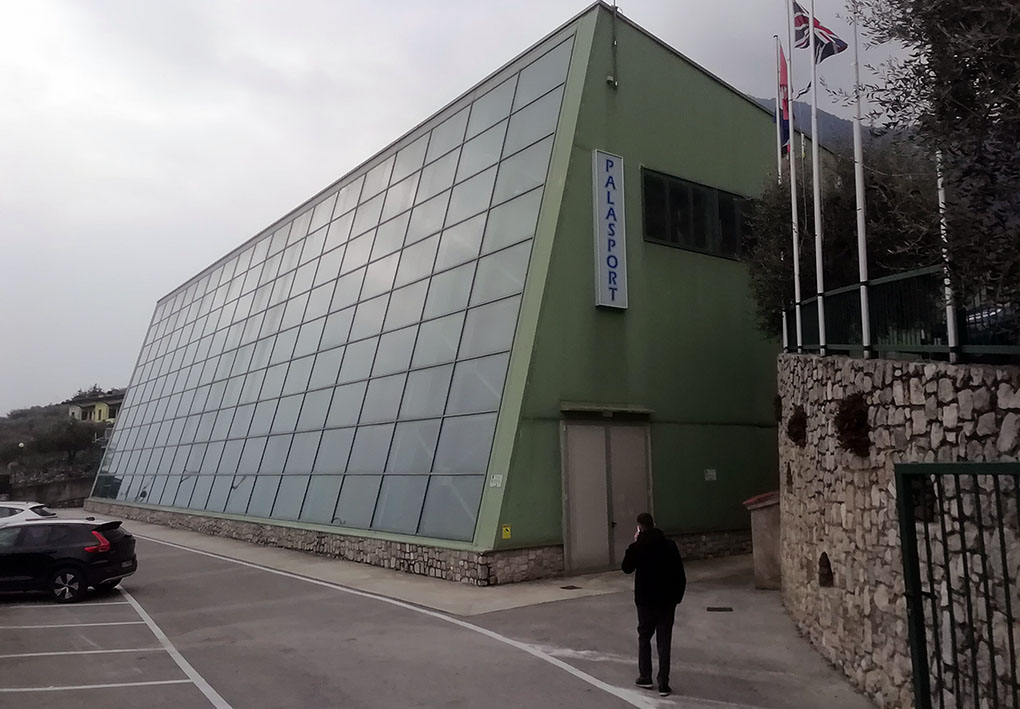 Edificio moderno con una struttura triangolare inclinata, caratterizzato da grandi pannelli di vetro rettangolari sulla facciata. Sul lato destro, un'insegna verticale blu con la scritta "PALASPORT" identifica l'edificio.