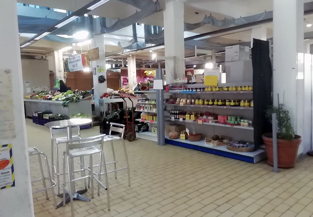 Interno di un mercato coperto con banchi di frutta e verdura fresca. A sinistra, tavoli e sedie in metallo per i clienti. A destra, scaffali con prodotti alimentari confezionati.