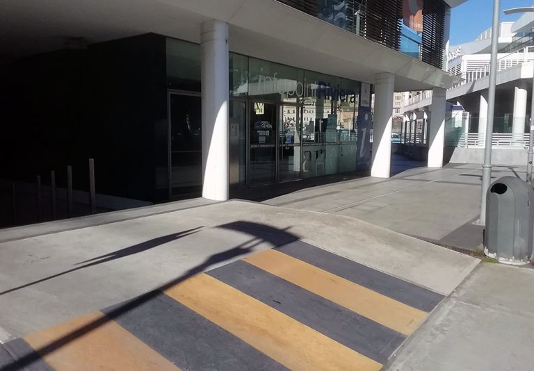 Ingresso di un edificio moderno con una facciata in vetro, sorretto da colonne bianche. Davanti all’entrata, un attraversamento pedonale con strisce gialle e nere. Sul lato destro, un cestino per i rifiuti e una parte di una passerella sopraelevata. Sullo sfondo, attraverso le vetrate, si intravedono edifici urbani.