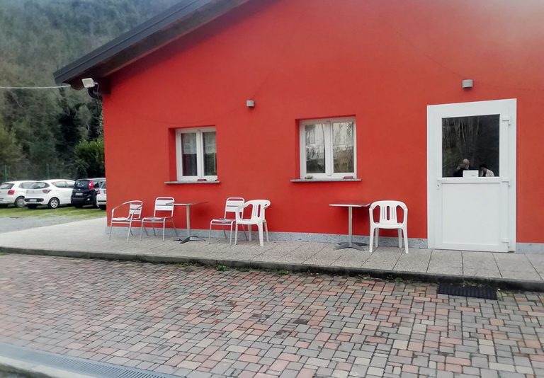 Edificio con parete arancione, due finestre e una porta bianca. Davanti, tre tavoli con sedie di plastica bianca su pavimento misto di cemento e mattoni. Sullo sfondo, auto parcheggiate e alberi.
