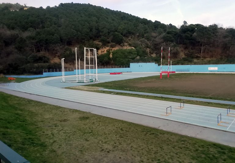 Pista di atletica azzurra con corsie segnate. Ostacoli posizionati lungo il percorso. Area centrale con attrezzature per il salto con l'asta. Zona erbosa e campo in terra adiacenti. Sfondo con una collina alberat