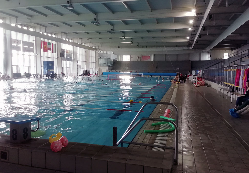 Piscina coperta con corsie delimitate da galleggianti. Alcune persone stanno nuotando. L'ambiente è illuminato da ampie finestre. Sulla destra, panchine con asciugamani e attrezzature da nuoto