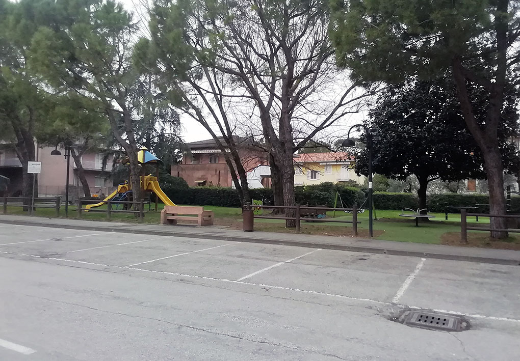 vista di un parcheggio con in lontananza la vista di un parcogiochi attrezzato per bambini in un prato, circondato da alberi
