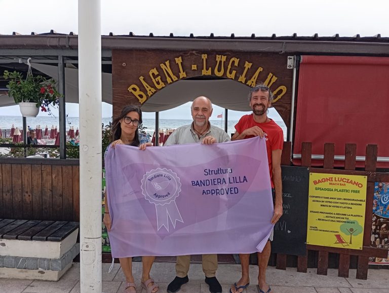 Davanti allo stabilimento dei Bagni Luciano, i proprietari posano sorridenti con il presidente di Bandiera Lilla. I tre mostrano la bandiera con il marchio Bandiera Lilla Approved