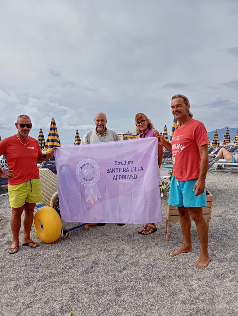 Il presidente di Bandiera Lilla posa sulla spiaggia con la proprietaria dei Bagni Lido e due bagnini dello stabilimento, mostrando la Bandiera con il marchio Bandiera Lilla Approved. Poco lontano da loro, una sedia JOB
