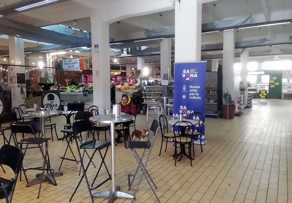 Mercato coperto con tavoli e sedie per la ristorazione. Stalli di frutta e verdura sullo sfondo.