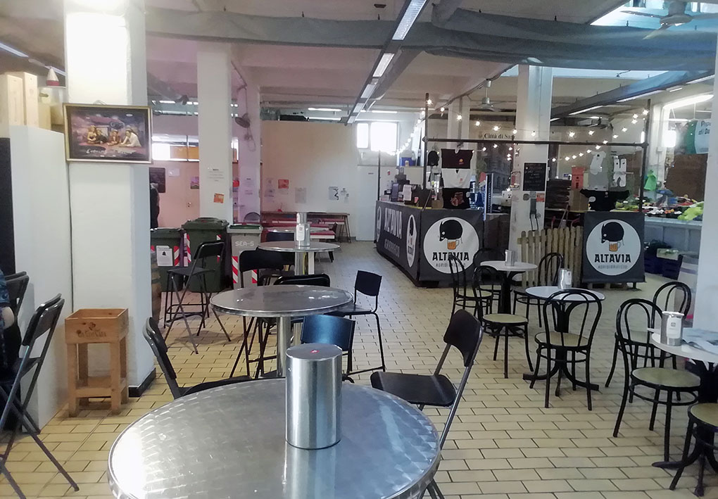 Spazio interno con tavoli e sedie metalliche nere. Bancone con insegna "ALTAVIA". Led luminosi al soffitto. Pavimento con piastrelle chiare e luce naturale dalle finestre.