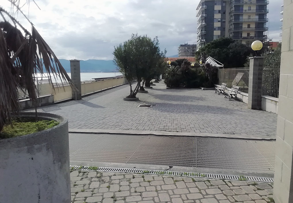 Passeggiata pavimentata lungo il lungomare, delimitata da una ringhiera sulla sinistra e alberi sulla destra. Panchine posizionate lungo il percorso. Sullo sfondo edifici.
