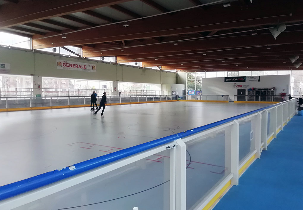 Pista di pattinaggio al coperto con pavimento chiaro e linee rosse per attività sportive. Barriera trasparente con bordo blu.