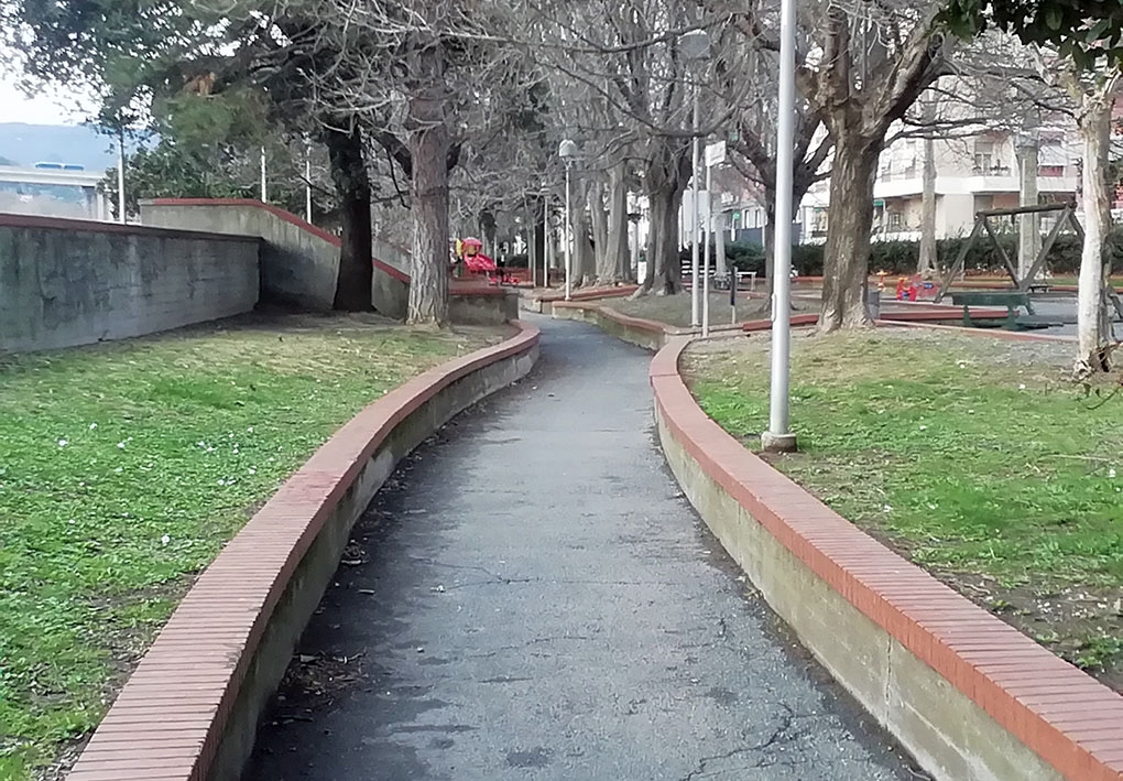 Sentiero pavimentato in un parco, delimitato da bassi muri in mattoni. Alberi spogli lungo il percorso, lampioni sulla destra e un’area giochi sullo sfondo