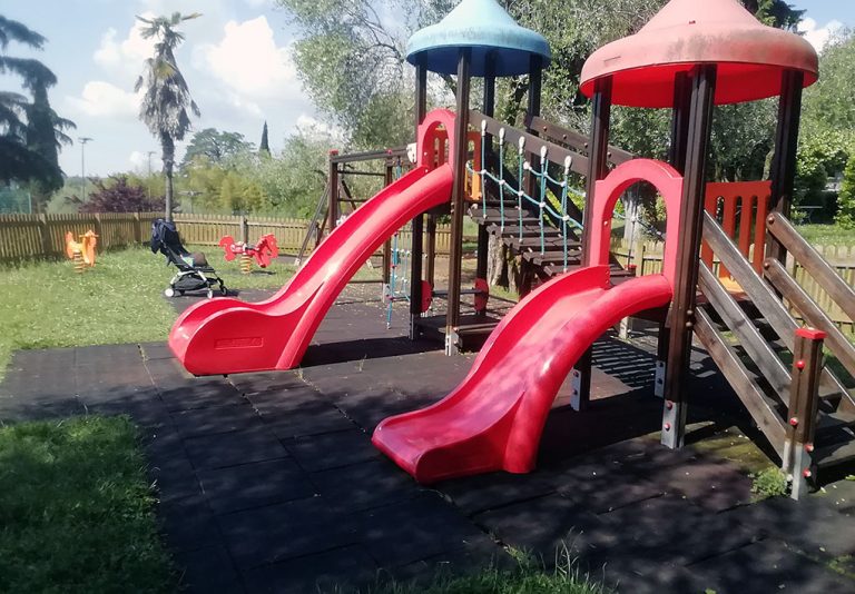 Parco giochi con in primo piano uno scivolo doppio e come sfondo dei giochi a molla. La pavimentazione è erbosa e antitrauma. Come sfondo alberi.
