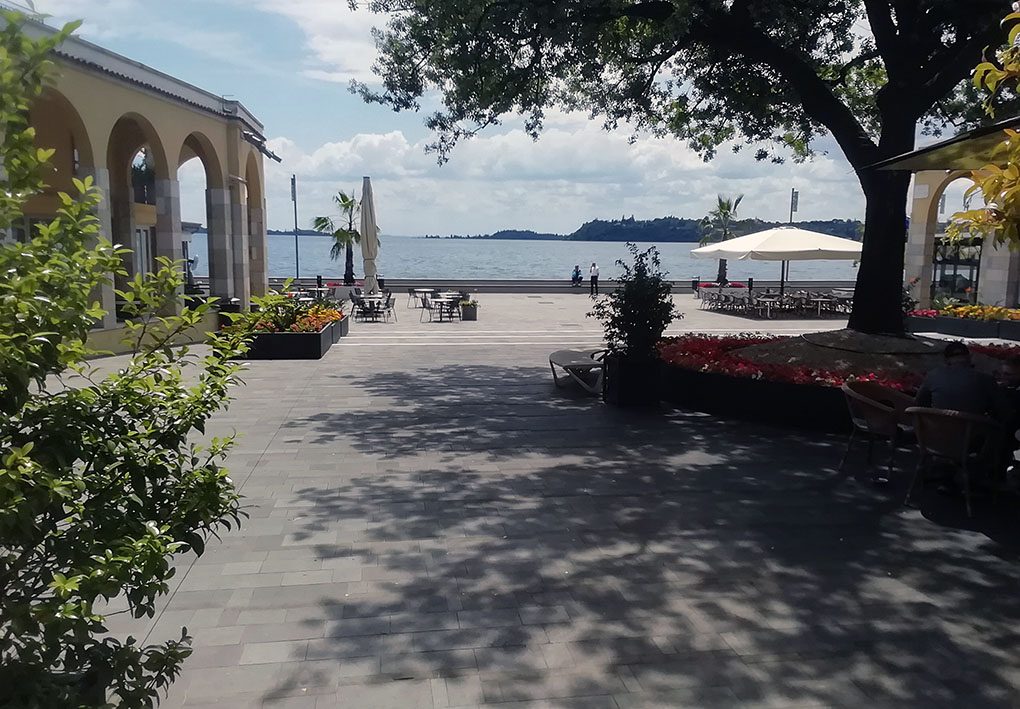 Zona pedonale lungo lago di Gardone Riviera con pavimentazione in lastricati di pietra, presenza di alberi e diversi edifici lungo la via.