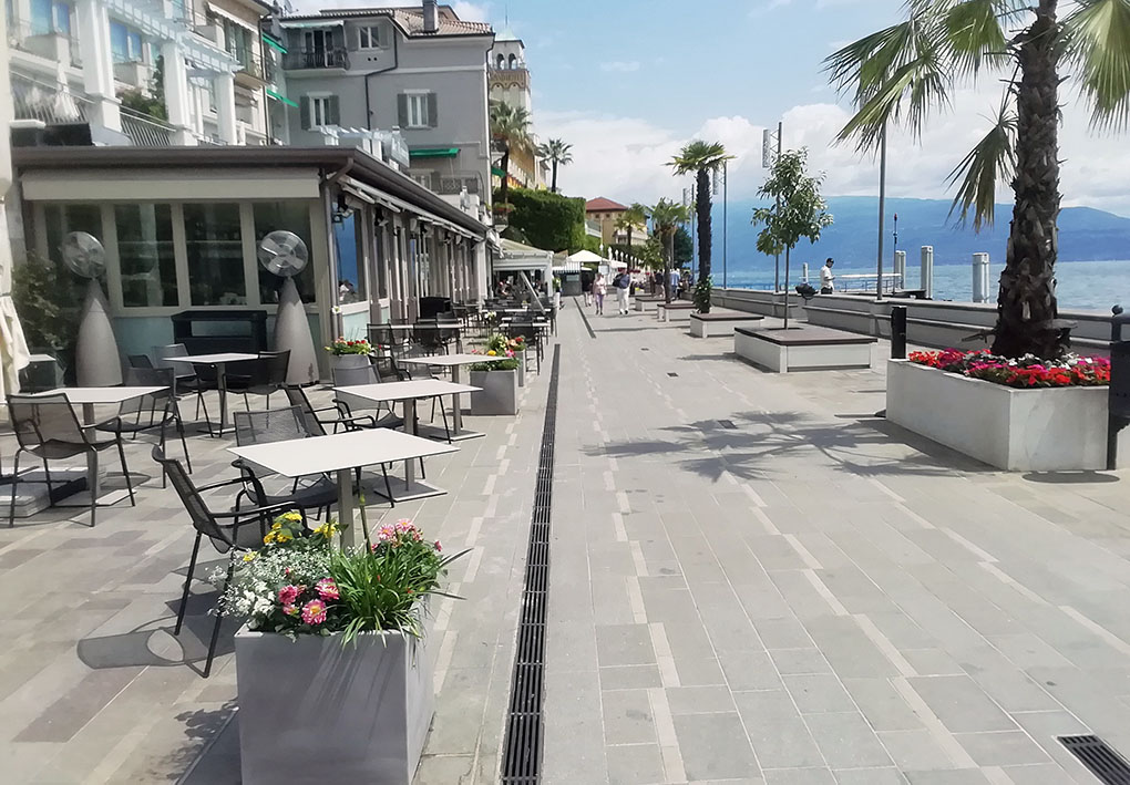 Lungolago pedonale con pavimentazione rugosa e senza dislivelli con alberi e locali. Come sfondo il lago e un limpido cielo