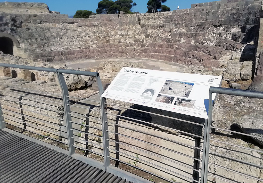 Passerella in legno con corrimano in metallo che affaccia su un anfiteatro romano in pietra. In primo piano, un pannello informativo con testo e immagini. Sullo sfondo, alberi.