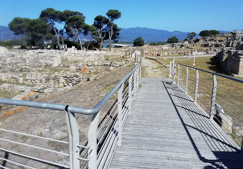 Passerella in legno con corrimano in metallo che attraversa il parco archeologico di Nora con resti di mura in pietra. Sullo sfondo, alberi e montagne.