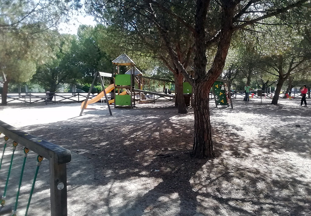 Parco giochi all'aperto con scivolo arancione, struttura in legno e pannelli verdi. Sullo sfondo, altalene, giochi a molla e alberi che offrono ombra.
