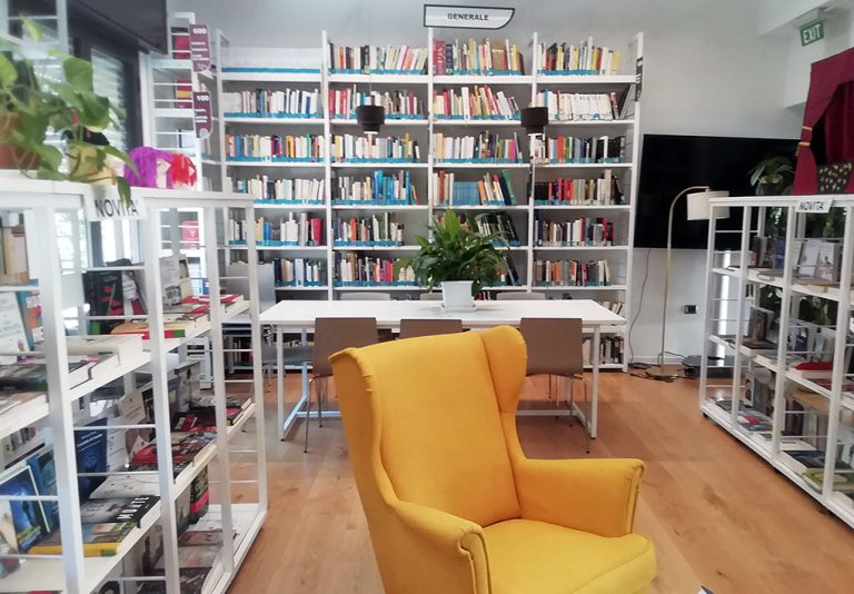 Biblioteca luminosa con scaffali bianchi pieni di libri. Al centro, un tavolo bianco con sedie e una pianta decorativa. In primo piano, una poltrona gialla. Sullo sfondo, una TV e un'uscita di emergenza.