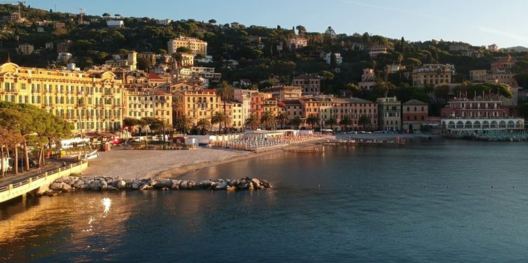 la spiaggia di ghiaia, ripresa da lontano, con alle spalle lo storico fronte a mare del borgo, composto da case colorate e storici alberghi di lusso. Alle spalle la collina ricca di alberi.