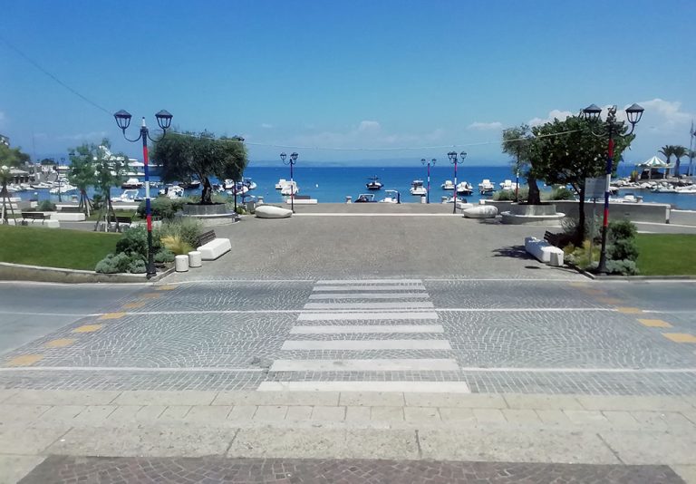 Attraversamento pedonale senza barriere architettoniche attraversa la pista ciclopedonale e conduce a un ampio marciapiede sul lungomare con pavimentazione liscia e senza ostacoli, fiancheggiato da alberi, panchine accessibili e lampioni. Sullo sfondo si apre un porto turistico con barche ormeggiate sotto un cielo terso, offrendo un ambiente calmo e invitante.