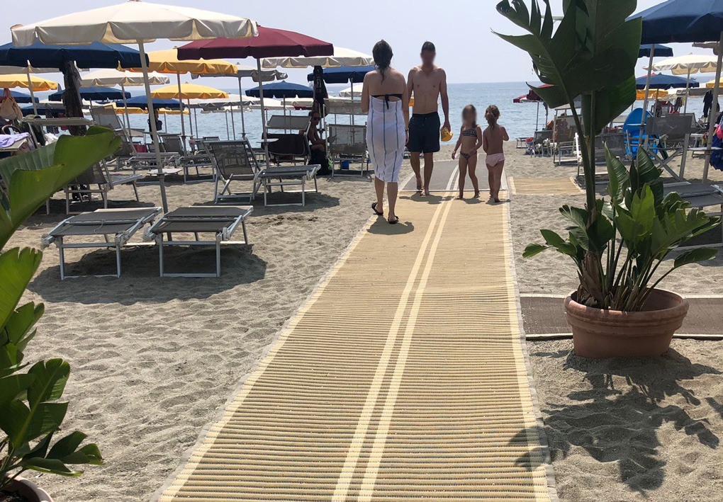 La passerella si allunga verso il mare, piante decorative abbelliscono l'entrata, la passerella si sviluppa verso il mare, tra ombrelloni ordinati su un fondale sabbioso. Una famiglia percorre la passerella.