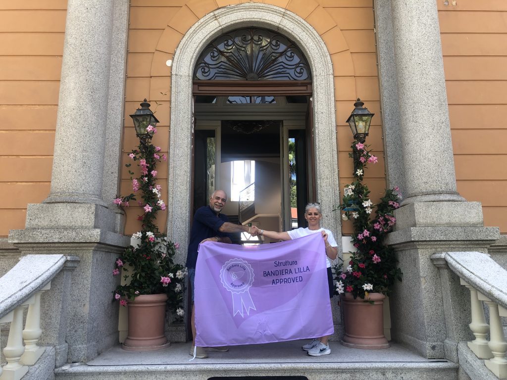 Il presidente di Bandiera Lilla posa con la proprietaria dell'Hotel Villa San Giuseppe e la bandiera "Bandiera Lilla Approved", davanti all'ingresso dell'albergo