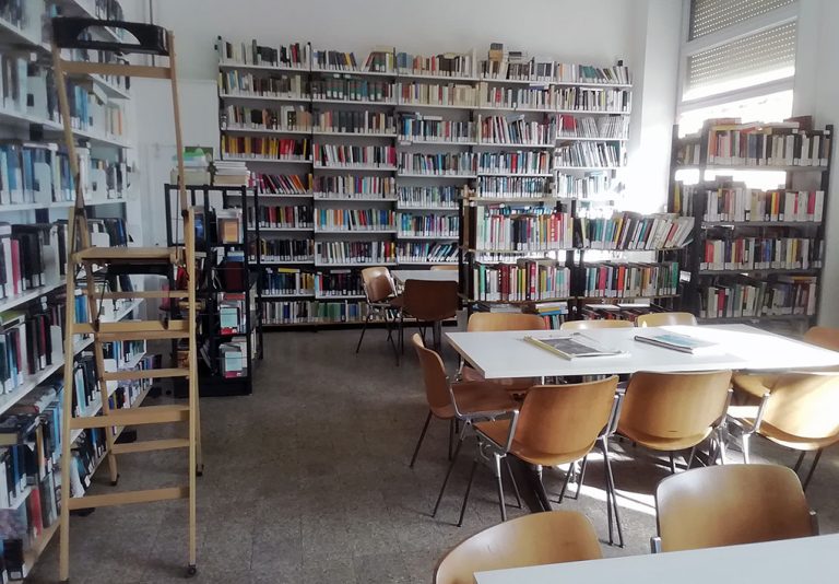 Interno di biblioteca con scaffali pieni di libri, tavoli e sedie al centro, scala di servizio contro le mensole.