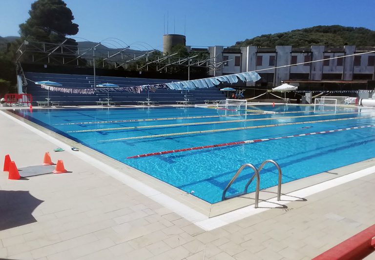 Piscina all’aperto con corsie delimitate da boe, bordo piastrellato con coni arancioni, gradini d’accesso sul lato destro, tribune coperte e edificio sullo sfondo circondato da alberi e colline.
